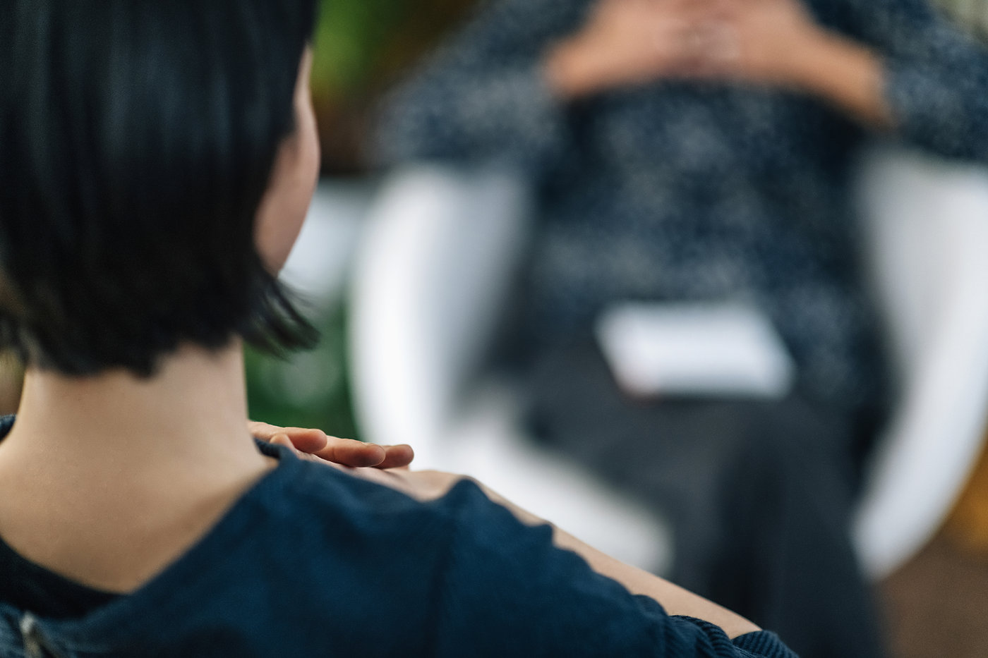 Person with short dark hair sits facing another person, blurred in the background, suggesting a discussion. Calm, muted colors.